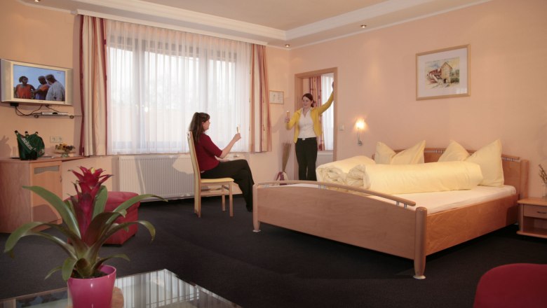 Doppelzimmer, © Fam. Graf Zwei Frauen in einem Hotelzimmer mit Doppelbett, eine sitzt, die andere steht mit erhobenem Glas.