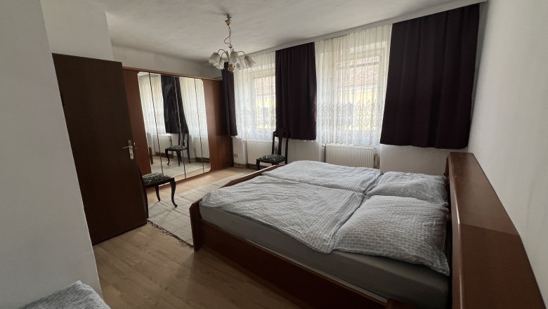 Doppelzimmer, © Gästehaus Weinwurm Schlafzimmer mit Doppelbett, Kleiderschrank mit Spiegel, Stuhl und Vorhängen an den Fenstern.