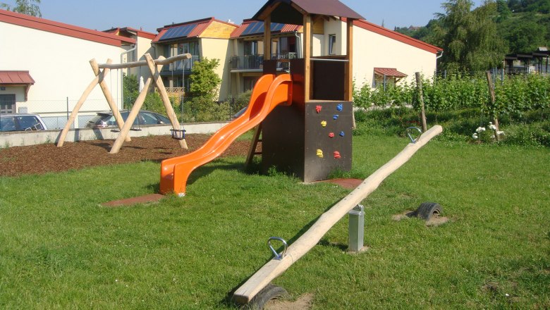 Kinderspielplatz, © Weingut Deutsch Ein Kinderspielplatz mit Rutsche, Schaukel und Wippe auf einer Wiese vor Wohnhäusern.