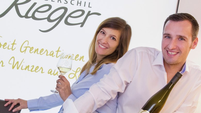 Wine tavern Oberschil-Rieger, © Oberschil-Rieger Two people are smiling and holding bottles of wine in front of a sign that reads 'Oberschil Rieger'.