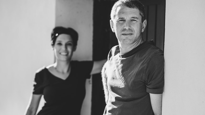 Willkommen!, © Michael Reidinger Schwarz-weiß Foto von zwei Personen mit Weinflaschen vor einer Tür.