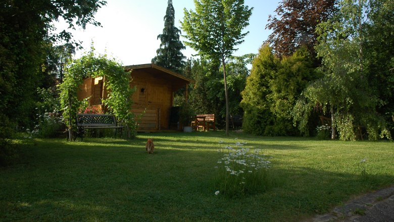 Garten zum Entspannen, © Christine Nigl Ein ruhiger Garten mit einer Holzhütte, Bäumen und einer Bank. Eine Katze läuft über den Rasen.