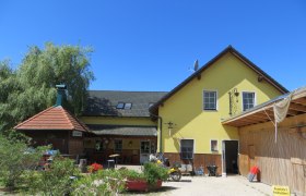 Gasthof Oase am Teich, © Urani Gelbes Gebäude mit grünem Dach und Terrasse, umgeben von Pflanzen, unter blauem Himmel.