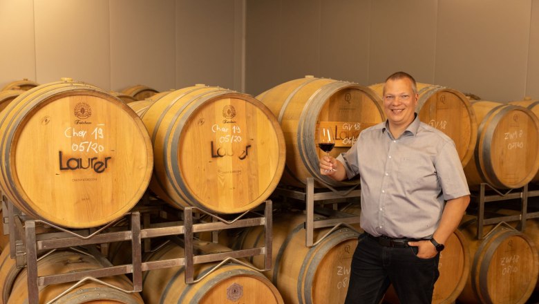 Barrel cellar, © Helge Woell Man smiling in front of wine barrels and holding a glass of red wine.