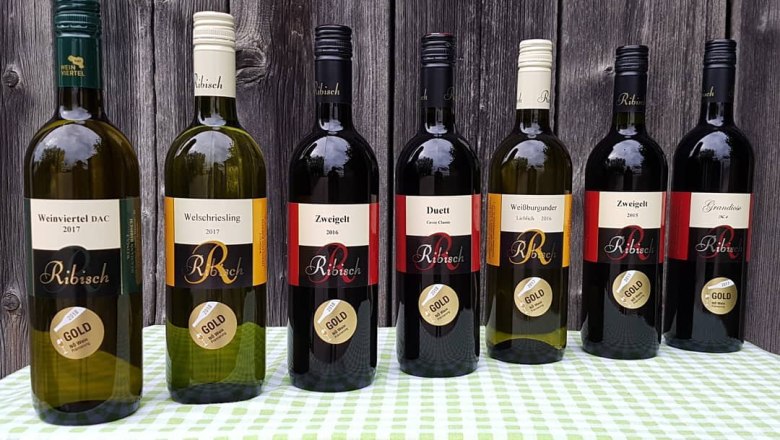 Weinsortiment, © Weingut Ribisch Sieben Weinflaschen der Marke Ribisch auf einem Tisch vor Holzwand.