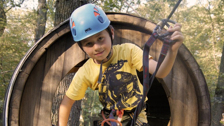 Kletterpark, © Fam. Freudhofmaier Ein Kind mit Helm und Kletterausrüstung in einem hölzernen Kletterpark-Tunnel.