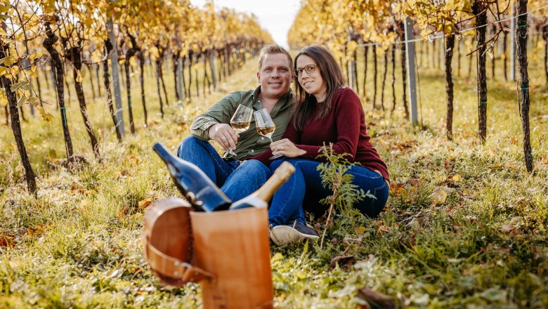 Rieder family, © Claudia Müllner A couple is sitting in a vineyard holding glasses of wine. In front of them is a picnic basket with a bottle of wine.