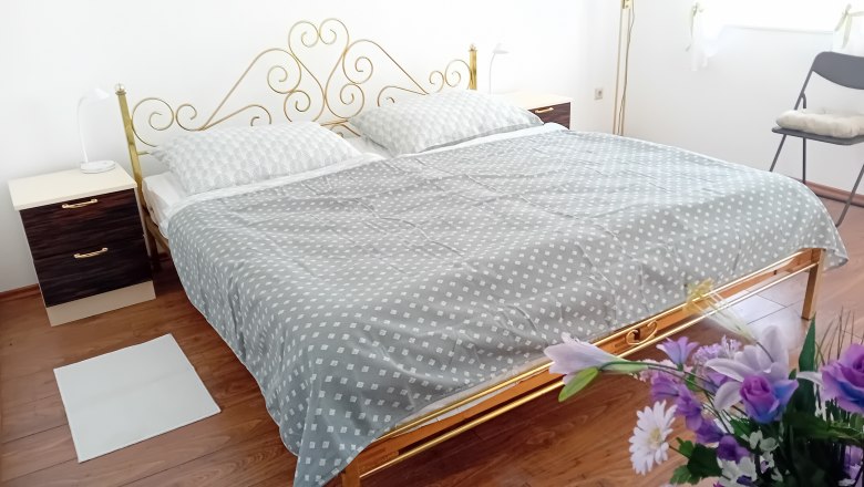 Schlafzimmer mit Doppelbett, © Erna Mitsch Schlafzimmer mit einem Doppelbett, goldfarbenem Kopfteil, zwei Nachttischen und Blumen im Vordergrund.