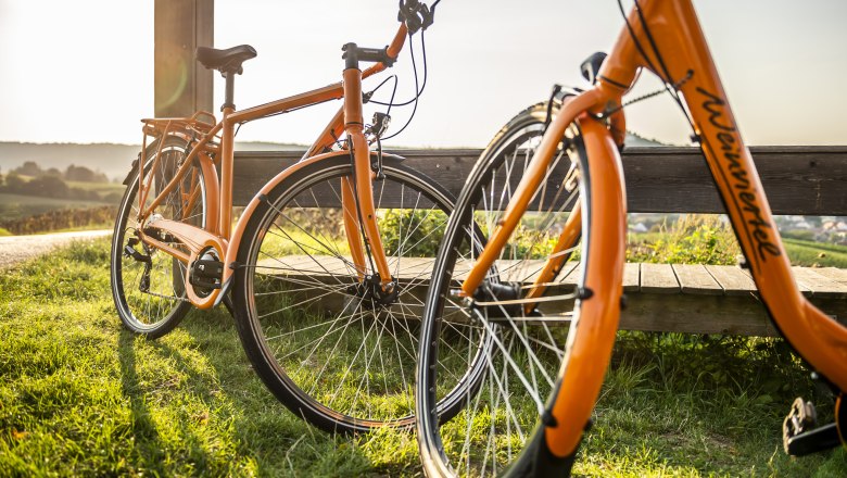 Radfahren im Weinviertel, © Weinviertel Tourismus GmbH / POV / Robert Herbst Zwei orangefarbene Fahrräder stehen auf einer Wiese neben einer Holzbank, im Hintergrund eine hügelige Landschaft bei Sonnenuntergang.