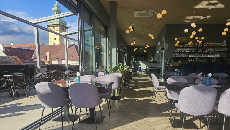 Rooftop Innenbereich, © Rebecca Freitag Innenbereich eines modernen Cafés mit Glaswänden, Blick auf den Rathausturm, Tische und Stühle, hölzerner Boden und dekorative Beleuchtung.