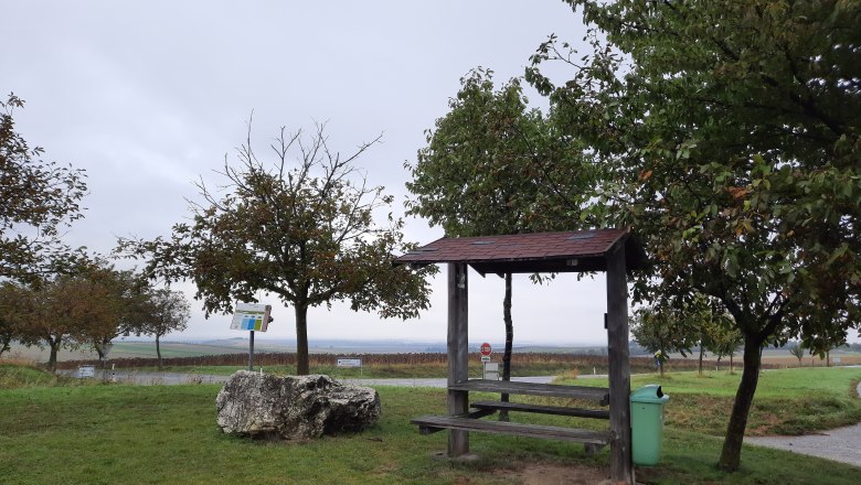 Rastplatz Siebenbergeblick, © Gemeinde Gaubitsch Ein Rastplatz mit Holzbänken und einem Tisch, umgeben von Bäumen und einer Wiese, unter einem bewölkten Himmel.