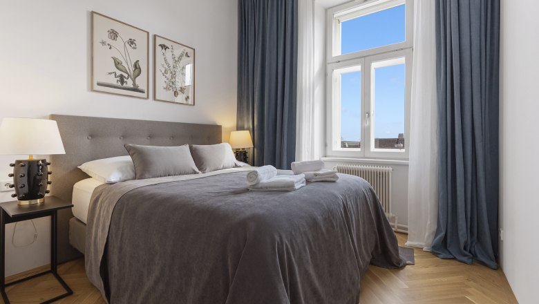 Gästezimmer, © Mt Ratzenboeck Ein elegantes Schlafzimmer mit einem großen Bett, grauer Bettwäsche, zwei Nachttischen mit Lampen und einem Fenster mit blauem Vorhang.