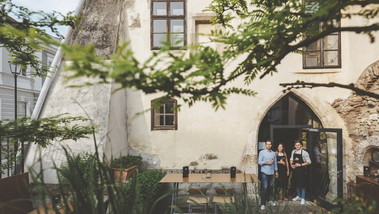 WeinStein guest garden, © Michael Reidinger Three people are standing in a historic courtyard in front of a building with a Gothic arched window.