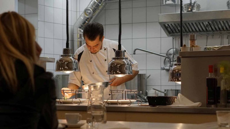 Christoph Schüller cooks in front of the guests, © Genusswirtschaft A chef in a white uniform prepares food in a restaurant kitchen while guests look on.