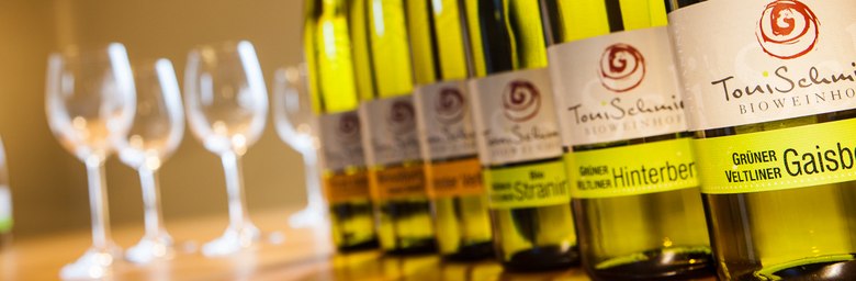 Weinflaschen, © Dieter Schewig Reihe von Weinflaschen mit Gläsern im Hintergrund.