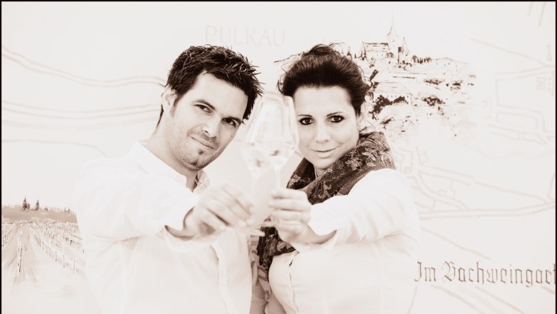 Simone und Hannes Jordan, © Petr Blaha Ein Mann und eine Frau stoßen mit Weingläsern an, im Hintergrund eine Zeichnung von Pulkau.