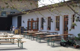 Buschenschank Demmer Innenhof, © Thomas Norek Innenhof einer Buschenschank mit leeren Holzbänken und einem Tisch, zwei Personen sitzen an einem Tisch, ein Spielzeugtraktor steht daneben.