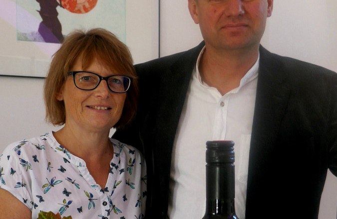 Familie Kugler, © Weingut Kugler Ein Mann und eine Frau stehen hinter einer Weinflasche des Weinguts Kugler. Im Vordergrund sind Weinblätter zu sehen.
