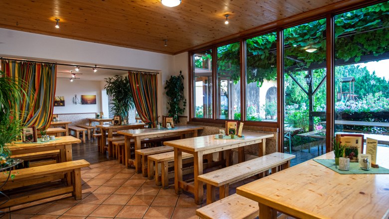 Heurigenlokal, © Martin Lifka Photography Innenansicht eines Heurigenlokals mit Holztischen und Bänken, bunten Vorhängen und großen Fenstern mit Blick auf einen begrünten Außenbereich.