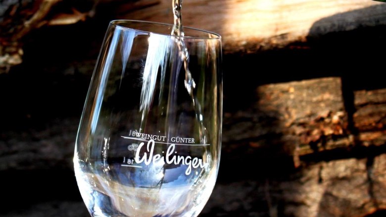 Weilinger Wein, © Weingut Günter Weilinger Ein Glas Wein wird aus einer Flasche mit orangefarbenem Verschluss eingeschenkt. Im Hintergrund sind Holzscheite zu sehen.