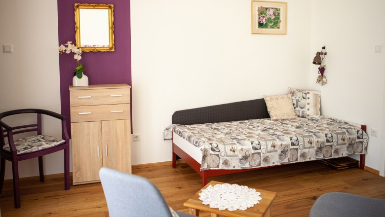 Schlafcouch, © Michael Sokolar Ein Wohnzimmer mit einer Schlafcouch, einem kleinen Schrank, einem Stuhl und einem Tisch auf Holzboden.