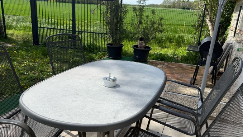 Sitzgelegenheit im Garten, © Dana Perl Gartentisch mit Stühlen auf einer Terrasse, umgeben von grünem Rasen und einem Zaun.