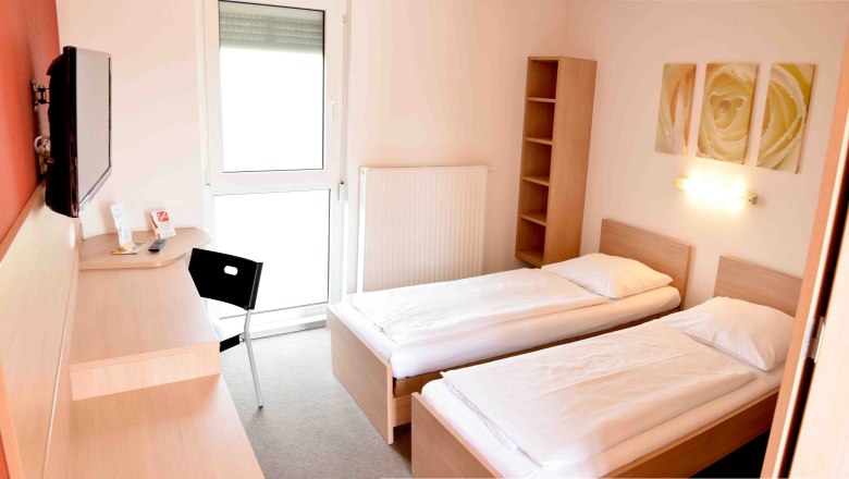 Motelzimmer, © Motel Angern Ein einfaches Motelzimmer mit zwei Einzelbetten, einem Schreibtisch, einem Stuhl und einem Fernseher an der Wand.