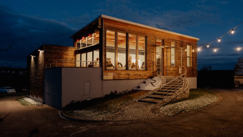 Wine tavern outside, © Weingut Rieder Exterior view of a modern wine tavern at night with illuminated windows and fairy lights.