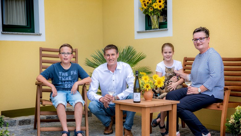 Familie Bauer, © R. Podolsky Eine Familie sitzt draußen auf Holzmöbeln vor einem gelben Haus. Ein Mann, eine Frau, ein Junge und ein Mädchen mit einer Katze sind zu sehen.