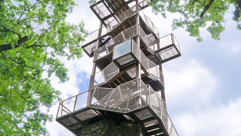 Kolisko card, © Johannes Zinser A modern observation tower made of metal and stone, surrounded by green trees, rises into the blue sky.