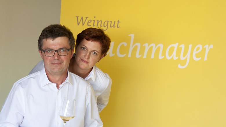 Maria und Thomas Buchmayer, © Michael Himml Zwei Personen in weißen Hemden vor gelbem Hintergrund mit der Aufschrift 'Weingut Buchmayer'. Eine Person hält ein Weinglas.