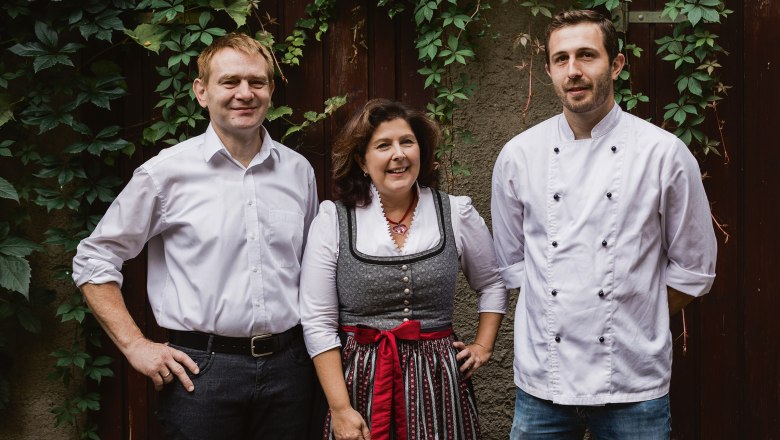 Host family: Christa, Stefan and Bernhard Fritsch, © Niederösterreich Werbung/David Schreiber Three people are standing in front of an ivy-covered wall, two men in white shirts and a woman in traditional dress.