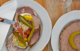 Heurigenjause, © Weinviertel Tourismus GmbH / Lisa Sedlatschek Zwei Teller mit belegten Broten und zwei Gläser Weißwein auf einem Holztisch.