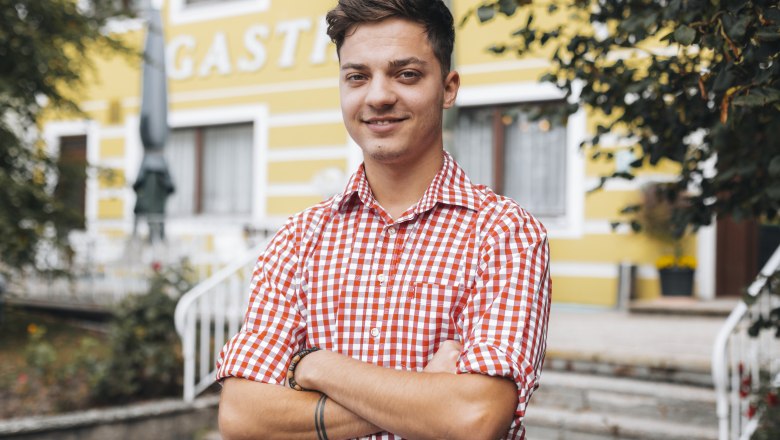 Im Service: Martin Scheiterer, © Niederösterreich Werbung/David Schreiber Ein junger Mann in kariertem Hemd steht lächelnd vor einem gelben Gebäude.