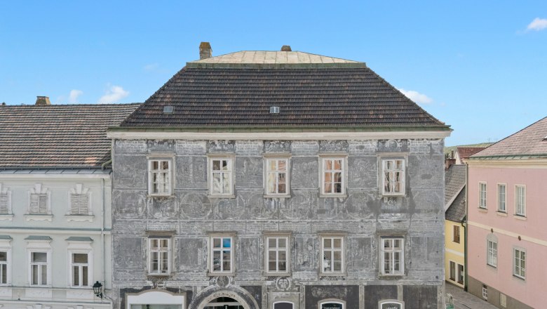 Herzlich Willkommen, © Mt Ratzenboeck Historisches Gebäude mit grauer Fassade und dekorativen Malereien in einer Stadt.