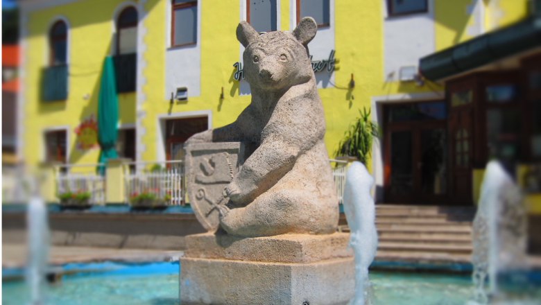 Matzen, © F. Brennig Steinbär-Skulptur mit Wappen in einem Brunnen vor einem gelben Gebäude.