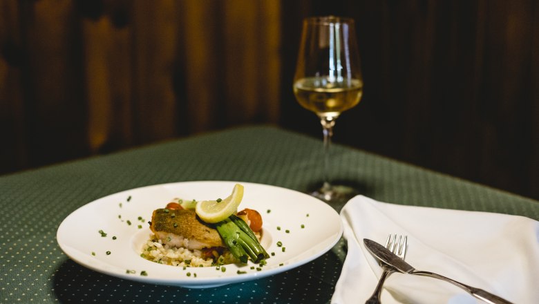 Lachsforelle mit Risotto, © Niederösterreich Werbung/David Schreiber Ein Teller mit Lachsforelle und Risotto, garniert mit Zitrone und Gemüse, steht auf einem gedeckten Tisch neben einem Glas Weißwein.