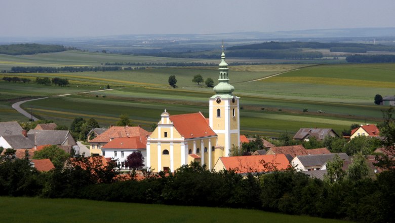 Ottenthal, © Gemeinde Ottenthal Blick auf eine Kirche in Ottenthal mit umliegenden Häusern und weiten Feldern im Hintergrund.