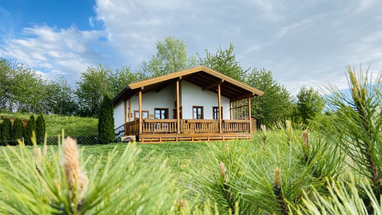 A quiet, relaxing place, © Weinstraße Weinviertel A small wooden house with a veranda stands on a green meadow, surrounded by trees and bushes under a cloudy sky.