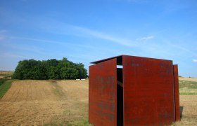Windwürfelhaus, © StadtGemeinde Mistelbach Rostiger Metallwürfel auf einem Feld mit Bäumen im Hintergrund.