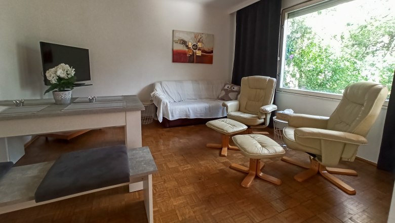 Wohn- und Essbereich, © Multimedia Lauda Wohnzimmer mit Sofa, zwei Sesseln, Tisch und großem Fenster.