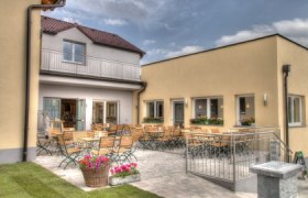 Heurigenlokal, © Fam. Falk Ein gemütlicher Außenbereich mit Holztischen und -stühlen vor einem gelben Gebäude.