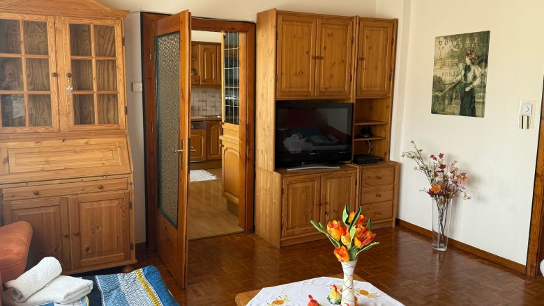 Wohnzimmer, © Dana Perl Wohnzimmer mit Holzmöbeln, einem Tisch mit Blumen und einem Fernseher.