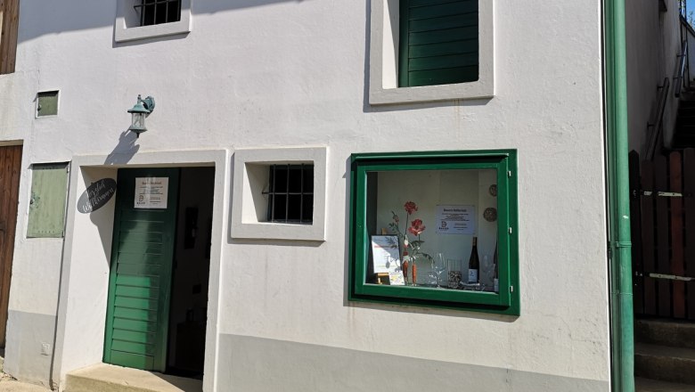 Bauers Kellerrast, © Weinbau Bauer Weiße Hausfassade mit grüner Tür und Fensterrahmen. Ein Schild mit der Aufschrift 'Bauers Kellerrast' ist sichtbar.