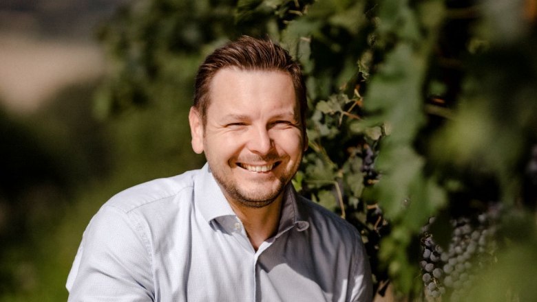 Weingut Heinisch, © Astrid Bartl Ein Mann in einem Weinberg, lächelnd und in die Hocke gehend, trägt ein helles Hemd und Jeans.