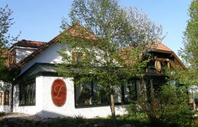 Kellertriftheuriger Loiskandl Aussenansicht, © Pfeifer Außenansicht eines traditionellen Gebäudes mit roten Dachziegeln und einem großen L-Logo an der Wand, umgeben von Bäumen und Pflanzen.