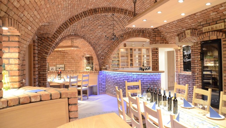 WeinWohnZimmer, © Weingut Wimberger Gemütlicher Weinkeller mit Backsteinwänden, gedecktem Tisch und Weinflaschen.