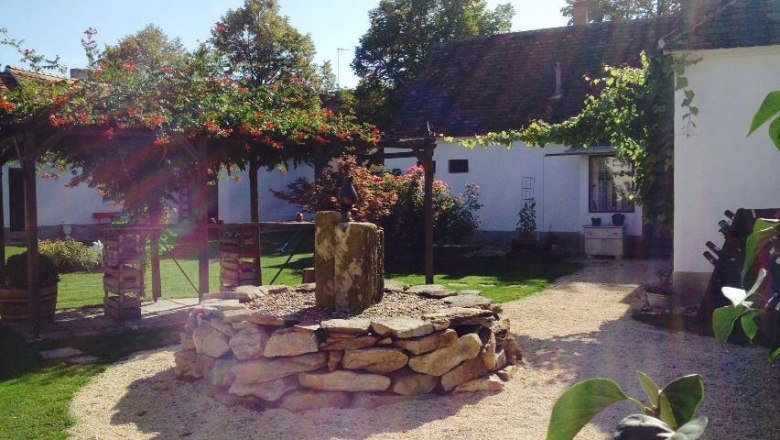 Garten, © Pollak Ein sonniger Garten mit einem Steinkreis und einem kleinen Brunnen in der Mitte, umgeben von weißen Gebäuden und grünen Pflanzen.