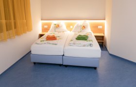 Doppelzimmer, © Sebastian Philipp Ein Doppelzimmer mit zwei Einzelbetten, gelben Vorhängen und blauer Boden. Die Betten sind mit Blumenbettwäsche und farbigen Handtüchern dekoriert.