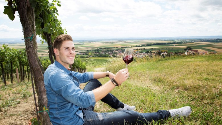 Patrick Kerner, © Weingut Kerner Ein Mann sitzt in einem Weinberg und hält ein Glas Rotwein. Im Hintergrund ist eine weite Landschaft zu sehen.